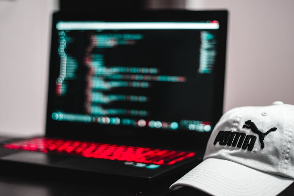 Close-up of a person's hand typing on a silver laptop keyboard, with the screen displaying lines of programming code. The laptop is placed on a wooden desk, and the person's wrist is adorned with a bl