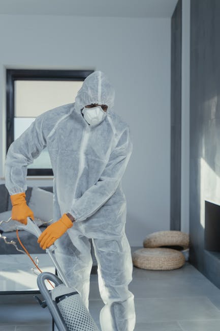 A professional cleaning technician dressed in a white protective suit, face mask, and blue gloves is performing deep cleaning or sanitisation of a carpet in a modern living room. The technician is usi